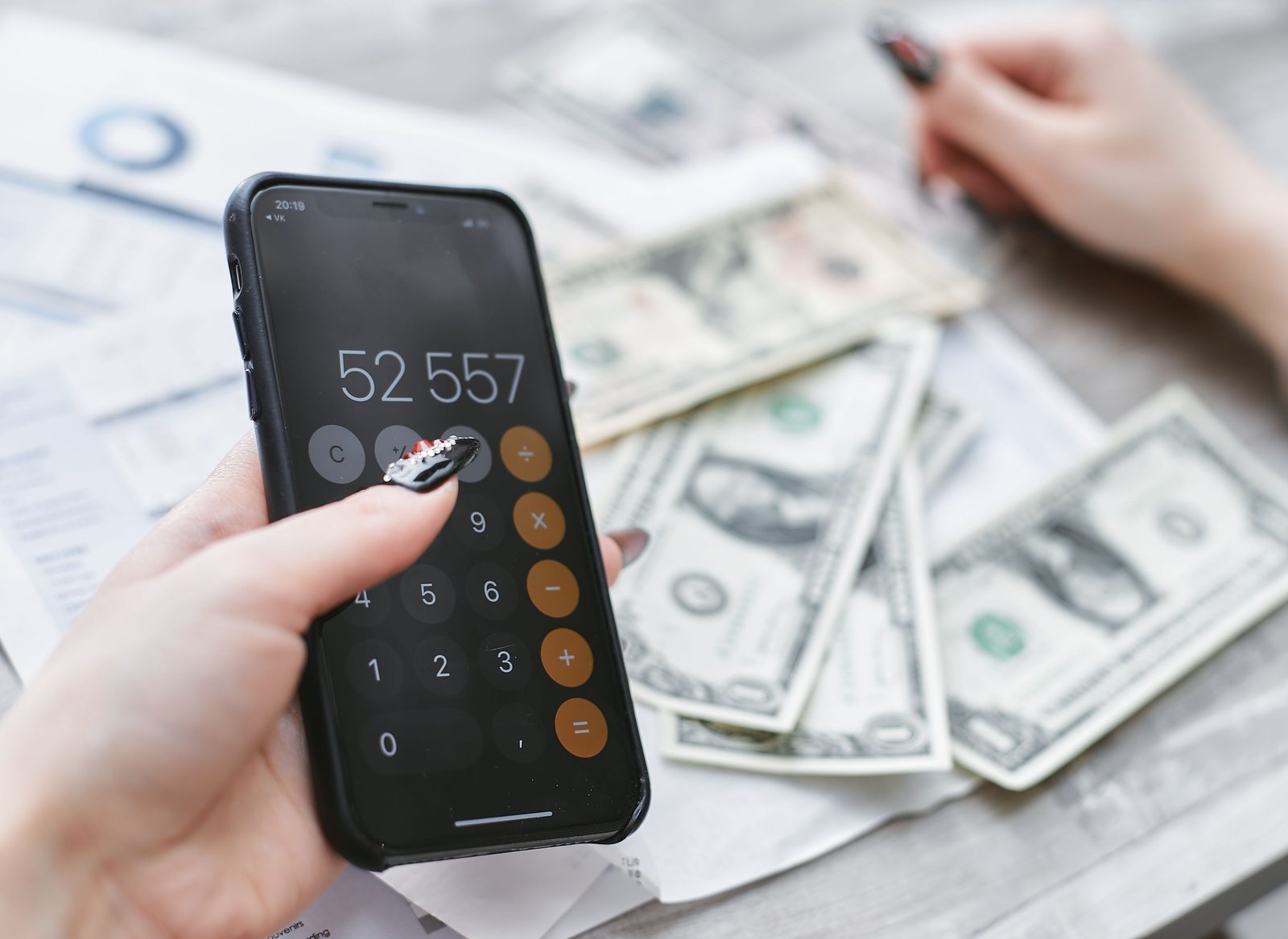 A hand using a phone calculator showing a large total next to scattered US dollar bills