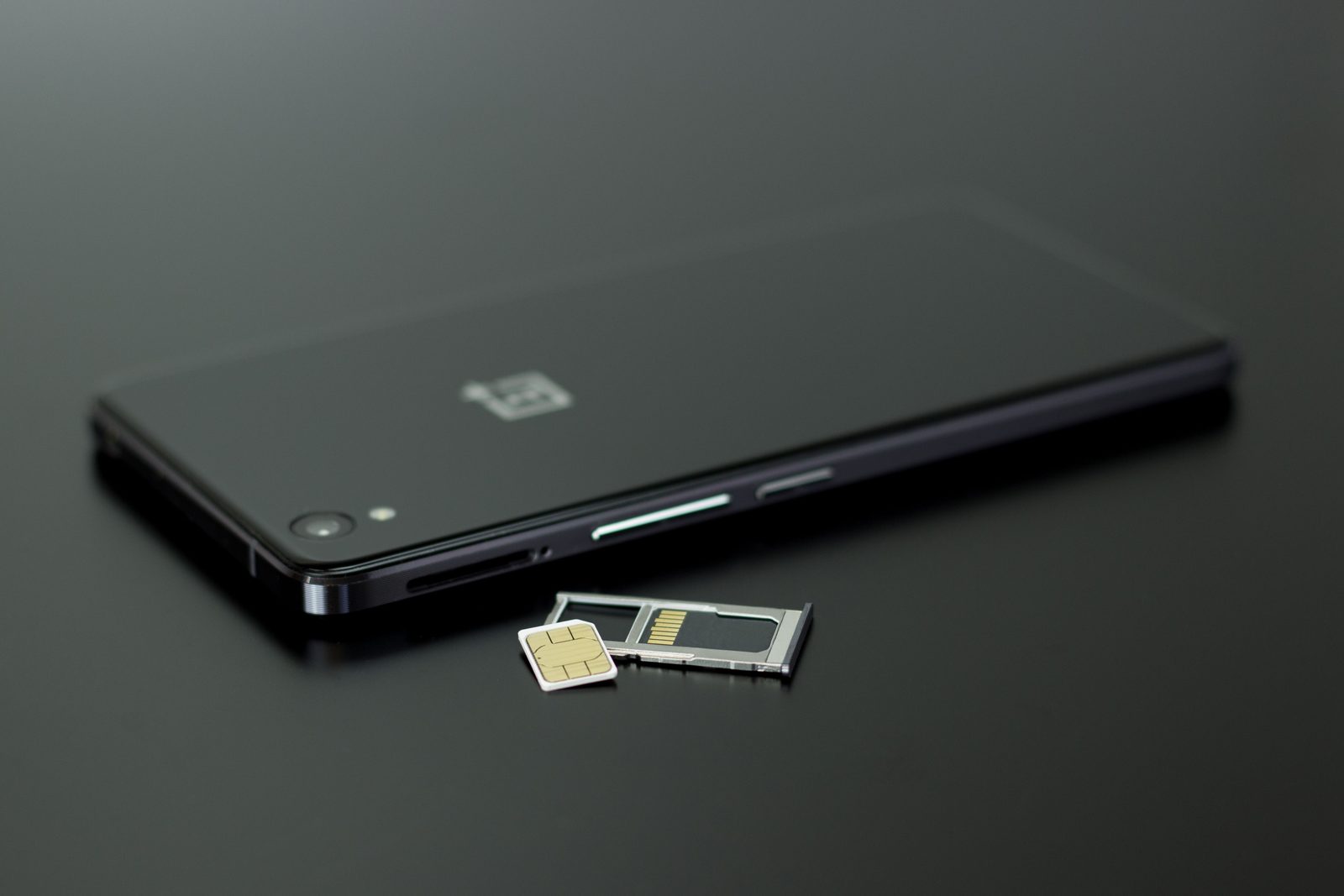 A black smartphone next to its removed SIM card and SIM tray on a dark surface